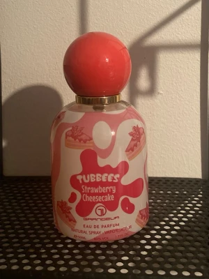 Tubbees Strawberry Cheesecake parfym - Parfymflaska med söt design inspirerad av jordgubbscheesecake. Flaskan är vit med rosa detaljer och har en stor rund rosa kork. Märket är Grandeur och doften heter Tubbees Strawberry Cheesecake. Perfekt för dig som gillar söta och lekfulla dofter.