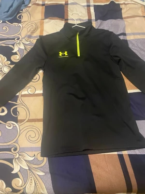 Svart Under Armour hoodie med dragkedja - Snygg svart hoodie från Under Armour med gul logga och dragkedja framtill. Tröjan har lång ärm och hög krage, perfekt för träning eller chill. Tillverkad i ett mjukt och stretchigt material som känns bekvämt mot huden.