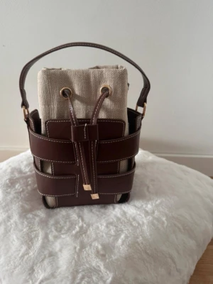 Brun och beige bucket handväska - Trendig bucket handväska i brun skinn med beige innerpåse i bomull. Väskan har snygga kontrastsömmar, justerbar axelrem och cool snörning upptill. Perfekt för dig som vill ha en unik accessoar med både stil och funktion.