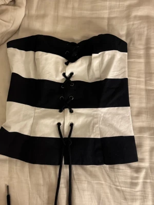 Randig korsettopp från H&M - Säljer en svartvit randig korsettopp från H&M med snörning framtill. Toppen är axelbandslös och har breda ränder i svart och vitt. Materialet känns som bomull och toppen har en snygg, figurnära passform.