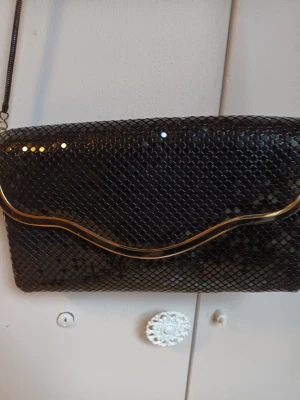 Svart glittrig kuvertväska med kedja - Snygg svart kuvertväska med glittrigt yttre och elegant guldfärgad vågig detalj på locket. Väskan har en tunn kedjerem som gör den enkel att bära över axeln. Perfekt accessoar för att lyfta din outfit.