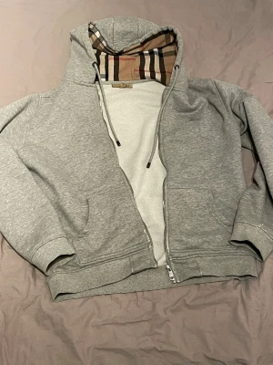 Grå zip hoodie från Burberry - Snygg grå zip hoodie från Burberry med klassiskt rutigt foder i huvan. Äkta gjord i italien. Pris diskuterbart vid snabb affär