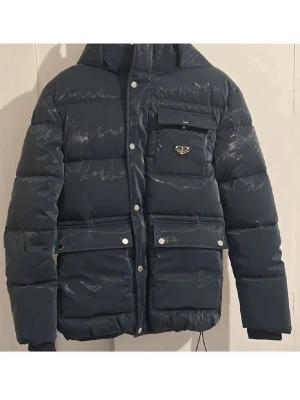 Mörkblå dunjacka från Philip plein - Kom privat för äktehetsbevis! Snygg mörkblå dunjacka från Philip plein med glansig finish, huva och flera fickor med tryckknappar. Jackan har en quiltad design och ett ikoniskt Prhilip plein emblem på bröstet. Perfekt för kalla dagar när du vill hålla stilen.