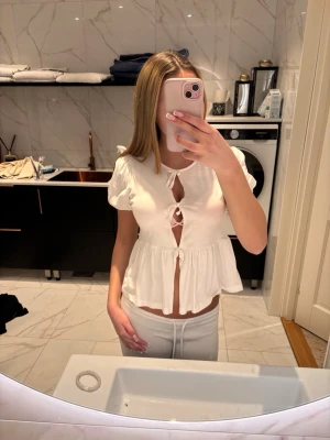 Vit blus med knyt och volang - Söt vit blus med korta puffärmar, knyt framtill och volang nedtill. Toppen har flera öppna detaljer längs bröstet och midjan, vilket ger en trendig look. Perfekt att styla med jeans eller kjol för en fräsch outfit.