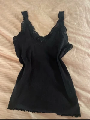 Svart spetskantad linnetopp - Snygg svart linnetopp med spetsdetaljer längs ringningen och axelbanden. Toppen har en v-ringad form och är ärmlös, perfekt att styla till olika outfits. Stretchigt material som sitter bekvämt och följer kroppen.