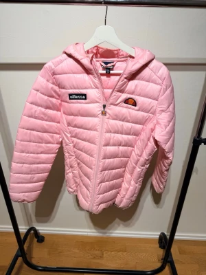 Rosa dunjacka från Ellesse med huva - Snygg ljusrosa dunjacka från Ellesse med huva och dragkedja framtill. Jackan har quiltad design och Ellesse-logga på bröstet. Perfekt för kyliga dagar och ger en sportig vibe till din outfit.
