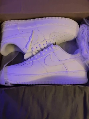 Nike Air Force 1 vita sneakers - Klassiska vita Nike Air Force 1 sneakers med låg profil och rund tå. Skorna har snörning, perforerade detaljer på tån och är tillverkade i skinn. Perfekta för dig som gillar en clean och tidlös stil.