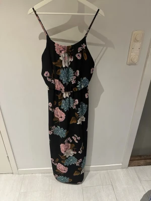 Blommig långklänning från ONLY storlek 36 - Svart långklänning från ONLY med tunna axelband och färgglatt blommönster i rosa, blått och brunt. Klänningen har en avslappnad passform med resår i midjan och är perfekt för varma dagar.