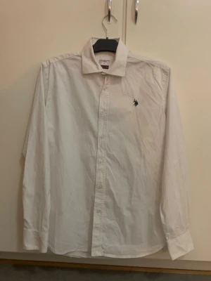 Vit skjorta från U.S. Polo Assn - Klassisk vit skjorta från U.S. Polo Assn i slim fit-modell. Skjortan har långa ärmar, knappar framtill och en liten broderad logga på bröstet. Tillverkad i bomull och passar perfekt till en clean och stilren look.