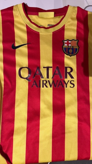 Barcelona matchtröja röd/gul Nike - Säljer en officiell Barcelona matchtröja från Nike med röda och gula vertikala ränder. Tröjan har klubbmärke på bröstet, Qatar Airways-tryck och La Liga-märke på ärmen. Perfekt för fotbollsfans som vill visa sitt stöd på läktaren eller planen.