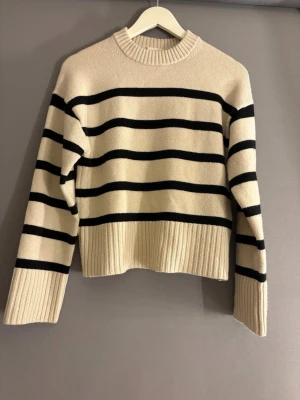 Beige stickad tröja med svarta ränder - Snygg beige stickad tröja med svarta horisontella ränder och ribbade muddar. Tröjan har rund halsringning och är något croppad i modellen. Perfekt för dig som gillar klassisk men trendig stil.