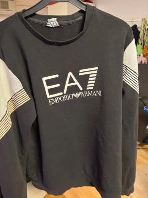 Svart sweatshirt EA7 Emporio Armani - Svart sweatshirt från EA7 Emporio Armani med vit logga framtill. Tröjan har kontrasterande ljusgrå partier och vita ränder på ärmarna. Rund halsringning och avslappnad passform. Perfekt för en sportig och trendig look.