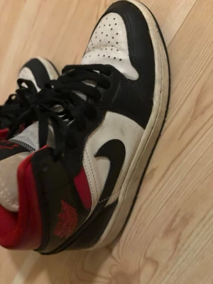 Nike Air Jordan 1 svart/vit/röd - Säljer ett par klassiska Nike Air Jordan 1 sneakers i svart, vitt och rött. Skorna har högt skaft, svart swoosh, perforerad tåbox och röda detaljer runt ankeln. Tillverkade i skinn med snörning och platt sula. Perfekta för dig som gillar ikoniska basketskor.