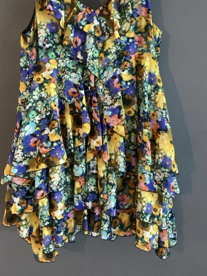 Blommig volangblus från the garden collection - Färgstark blommig blus från the garden collection med volanger och ärmlös design. Mönstret går i gult, blått, grönt och rosa. Luftigt material och v-ringad halsringning med flera lager volanger som ger en cool och lekfull look.