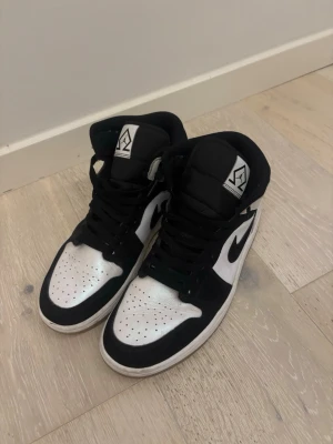 Nike Air Jordan 1 svart/vit sneakers - Snygga Nike Air Jordan 1 sneakers i svart och vitt med klassisk swoosh på sidan och Air Jordan-logga vid ankeln. Skorna har snörning, rund tå och platt sula. Perfekta för dig som gillar streetwear och vill ha en ikonisk look.