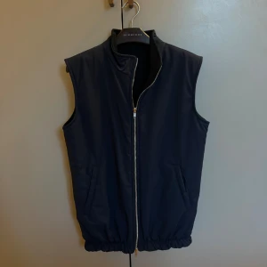 Mörkblå vändbar väst från L'Amoure - Stilren mörkblå vändbar väst från L'Amoure med hög krage och dragkedja framtill. Ena sidan är slät och blank, andra sidan är matt och mjuk. Två fickor på varje sida och elastisk nederkant för en snygg passform. Storlek S i nyskick.