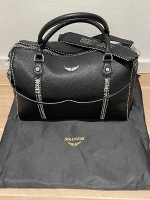 Svart handväska Zadig & Voltaire - Cool svart handväska från Zadig & Voltaire i skinn med silverfärgade detaljer och dragkedjor. Väskan har både handtag och axelrem, samt dekorativa nitar längs kanterna. Perfekt för dig som vill ha en edgy och stilren accessoar.