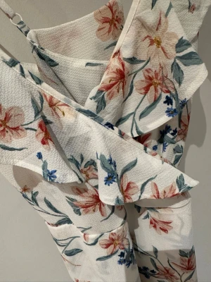 Blommig omlottklänning från H&M Divided - Säljer en vit omlottklänning från H&M Divided med blommigt mönster i rött, blått och grönt. Klänningen har tunna axelband och volangdetaljer längs kanterna. Perfekt för sommarens alla tillfällen!