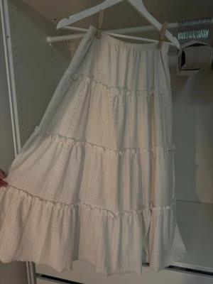 Vit långkjol med volanger - Säljer en romantisk vit långkjol med broderade blommor/hål och flera volanglager. Kjolen har resår i midjan för skön passform och är gjord i ett mjukt bomullstyg. Perfekt för dig som gillar en luftig och feminin stil. Gudomligt vacker, speciellt utomlands🥹 Den har lite missfärgningar högst upp vid linningen därav det billiga priset🤍