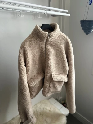 Beige teddyjacka med fickor - Mysig beige teddyjacka med hög krage och stora fickor framtill. Jackan har croppad passform, lång ärm och stängs med dragkedja. Perfekt för dig som vill ha en trendig och bekväm look till vardags.