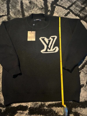 Svart stickad tröja med LV-logga - Svart stickad tröja från Louis Vuitton med stor vit LV-logga på bröstet. Tröjan har ribbstickad krage, ärmslut och nederkant. Klassisk passform och lång ärm. Perfekt för dig som gillar exklusiva och stilrena plagg.