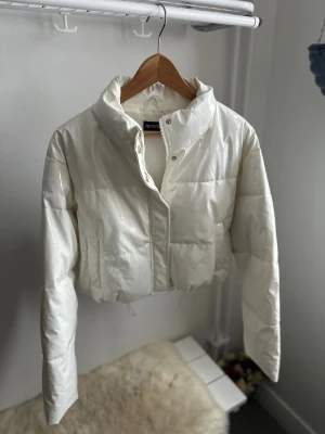 Vit croppad pufferjacka PrettyLittleThing - Snygg croppad pufferjacka från PrettyLittleThing i glansig vit färg. Jackan har hög krage, dragkedja och tryckknappar framtill. Perfekt för att ge din outfit en trendig vibe under kyliga dagar.