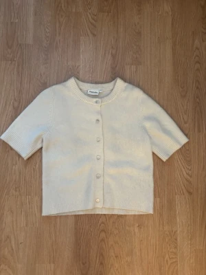 Krämvit kofta från Kappahl  - Kortärmad kofta i mjuk, krämvit stickad kvalitet från Kappahl. Rund halsringning, ribbade kanter och knäppning framtill med matchande knappar. Perfekt för lager på lager och enkel att styla till olika looks.