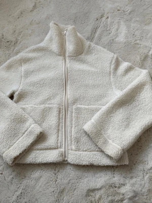 Beige teddyjacka med dragkedja - Mysig beige teddyjacka med hög krage och hel dragkedja framtill. Jackan har två stora fickor och är tillverkad i mjukt teddy-material som ger en fluffig och varm känsla. Perfekt för kyliga dagar och enkel att matcha med olika outfits.