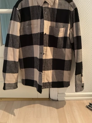 Rutig overshirt från H&M i beige och svart - Snygg overshirt från H&M i stor rutig design med beige och svarta rutor. Skjortan har klassisk krage, bröstficka och knäppning framtill. Perfekt för lager-på-lager och ger en avslappnad vibe till din outfit.