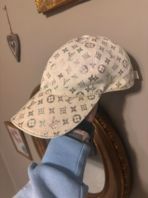 Beige Louis Vuitton keps med monogram - Snygg vit keps från Louis Vuitton med klassiskt monogrammönster i flera färger. Kepsen har böjd skärm och justerbar rem bak. Tillverkad i slitstarkt tyg som ger en exklusiv känsla och passar perfekt till streetwear eller en trendig look.