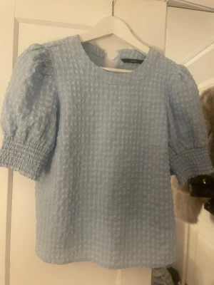 Ljusblå puffärmad blus - Supersöt ljusblå blus med puffiga korta ärmar och smockdetaljer vid ärmsluten. Blusen har ett strukturerat rutmönster i tyget och rak passform. Perfekt för dig som gillar en feminin och trendig look.