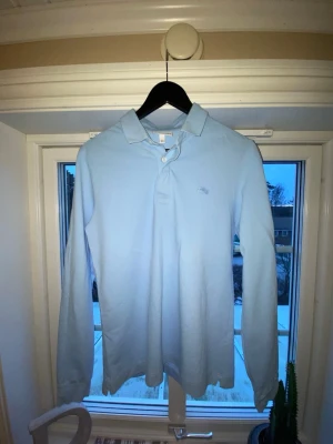 Ljusblå långärmad piké från Lacoste - Snygg ljusblå långärmad pikétröja från Lacoste med klassisk krage och knappar framtill. Tröjan har det ikoniska krokodilmärket broderat på bröstet och är tillverkad i mjuk bomull. Perfekt för en clean och stilren look.