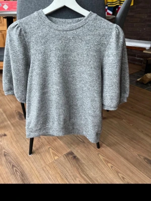 Grå puffärmad topp i bomull - Mysig grå topp med puffärmar och rund hals. Tillverkad i mjuk bomullsmix och har en enkel, stilren design som passar perfekt till jeans eller kjol. Puffärmarna ger en trendig touch och toppen har en normal passform.