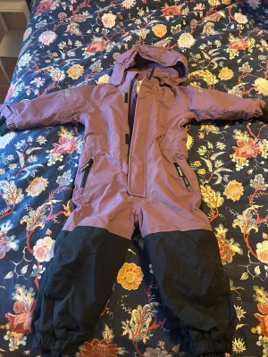 Lila och svart vinteroverall med huva - Overallmodell med lila överdel och svarta ben som enbart provats här hemma. Utrustad med huva, dragkedja framtill och reflexdetaljer på ärmar och rygg. Två fickor med dragkedja och resår vid benslut. Perfekt för kalla vinterdagar.