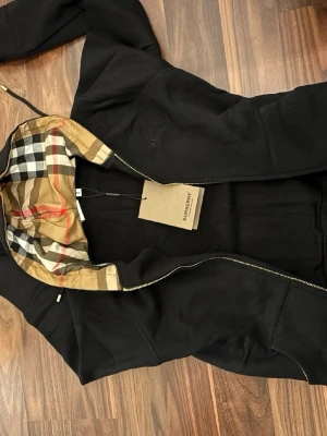 Svart vindjacka från Burberry - Snygg svart vindjacka från Burberry med klassiskt rutigt foder i huvan. Jackan har dragkedja framtill och en stilren look med diskret Burberry-logga på bröstet. Perfekt för dig som vill ha en ikonisk jacka med exklusiv känsla.