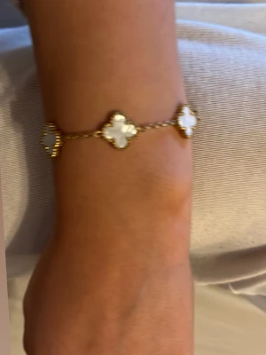 Guldarmband med vita fyrklöver - Snyggt armband från dressy i guldton med fem vita fyrklöverformade hängen, troligen i pärlemor. Kedjan är tunn och elegant, och varje klöver har en detaljerad kant. Perfekt accessoar för dig som gillar stilrena och trendiga smycken.