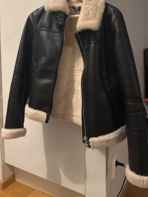 Svart skinnjacka med fuskpäls - Snygg svart skinnjacka med beige fuskpäls på krage, ärmslut och nederkant. Jackan har dragkedja framtill och är fodrad med mjuk pälsimitation för extra värme. Perfekt för kyliga dagar och ger en cool look.