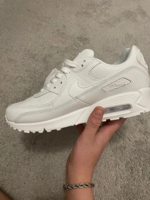 Nike Air Max 90 vita sneakers - Nike Air Max 90 sneakers i helvitt med klassisk siluett och synlig Air-enhet i sulan. Skorna har snörning, rund tå och är tillverkade i en mix av skinn och syntetmaterial. Perfekta för dig som gillar stilrena och tidlösa sneakers.