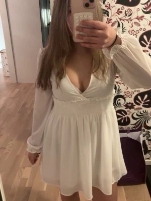 Vit omlottklänning med lång ärm - Säljer en vit enkel kortklänning med omlottdesign och v-ringning. Klänningen har långa, lätt puffiga ärmar och smock i midjan för en snygg passform. Tillverkad i ett tunt, flowy material som ger en luftig känsla. Perfekt för sommarens alla tillfällen!
