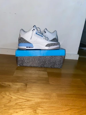 Air Jordan 3 White/Blue sneakers - Säljer ett par Air Jordan 3 sneakers i vitt läder med blå detaljer och den klassiska elephant print runt hälen och tån. Skorna har vita skosnören, blå inslag på sulan och öglor samt en bekväm, vadderad insida. Perfekta för dig som gillar streetwear och ikoniska modeller.