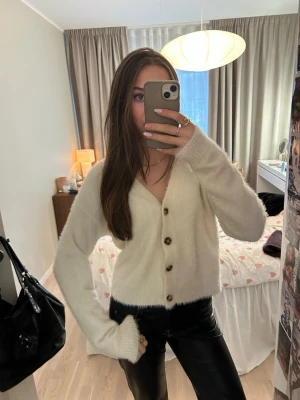 Vit fluffig kofta med knappar - Mysig vit kofta med v-ringning och stora bruna knappar framtill. Koftan är fluffig och har långa ärmar med ribbade muddar. Inga defekter!!! Tar emot bud!!!💞💞💞