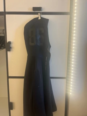 Svart hoodie med dragkedja och tryck - Säljer en svart hoodie med dragkedja och huva. På ryggen finns ett stort tryck med siffran 86 i svart. Hoodien har fickor framtill och snören i huvan. Perfekt för en avslappnad streetwear-look.