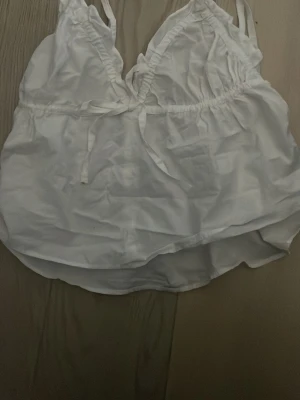 Vit linnetopp med knytband - Söt vit linnetopp med smala axelband och knytband framtill. Toppen har en luftig passform och lätt v-ringad halsringning. Perfekt för varma dagar och enkel att styla med jeans eller kjol.