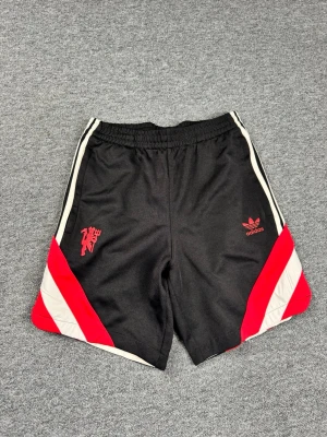 Adidas Manchester United shorts svart/röd - Svarta Adidas shorts med röda och vita detaljer samt Manchester United-logga på ena benet. Klassisk Adidas Trefoil-logga i rött på andra benet. Elastisk midja och sportig design, perfekta för fotboll eller träning.