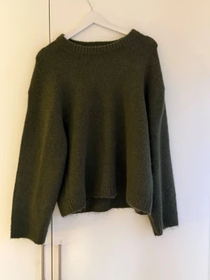 Olivgrön stickad oversized tröja - Mysig olivgrön stickad tröja med avslappnad passform och rund halsringning. Tröjan har långa ärmar och ribbstickade muddar vid ärmslut och nederkant. Perfekt för lager-på-lager och enkel att styla till olika outfits.