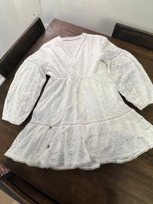 Vit broderad kortklänning med volang - Söt vit kortklänning i bomull med broderade detaljer och volangkant. Klänningen har v-ringning, långärmade puffärmar och fina spetsdetaljer längs kanterna. Prefekt till studenten