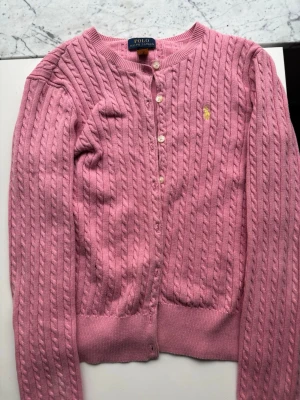 Rosa kabelstickad kofta Polo Ralph Lauren - Kabelstickad rosa kofta från Polo Ralph Lauren med rund halsringning och knappar framtill. Klassisk gul broderad logga.                                            Storlek 12-14 år. 