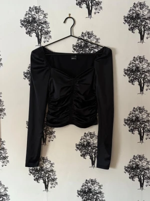 Svart långärmad croppad topp - Trendig svart långärmad topp med croppad passform och snygg rynkning framtill. Fyrkantig urringning och puffiga axlar ger en modern vibe. Tillverkad i ett glansigt syntetmaterial som känns mjukt mot huden. Perfekt för dig som vill sticka ut.