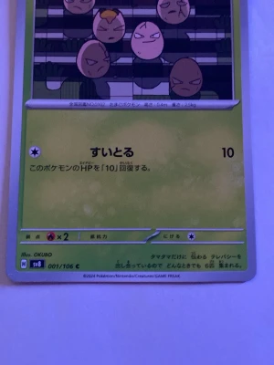 Japansk Pokémonkort Exeggcute - Japanskt Pokémonkort med Exeggcute-motiv från 2024. Kortet har grön bakgrund och japansk text, med illustration av sex äggliknande figurer. Kortnummer 001/106, illustratör OKUBO. Perfekt för samlare av Pokémonkort.