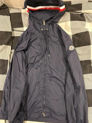 Marinblå vindjacka från Moncler - Snygg marinblå vindjacka från Moncler med huva och dragskedja. Jackan har dragkedja framtill, Moncler-logga på ärmen och rödvit rand på huvan. Perfekt för blåsiga dagar och enkel att matcha med det mesta.